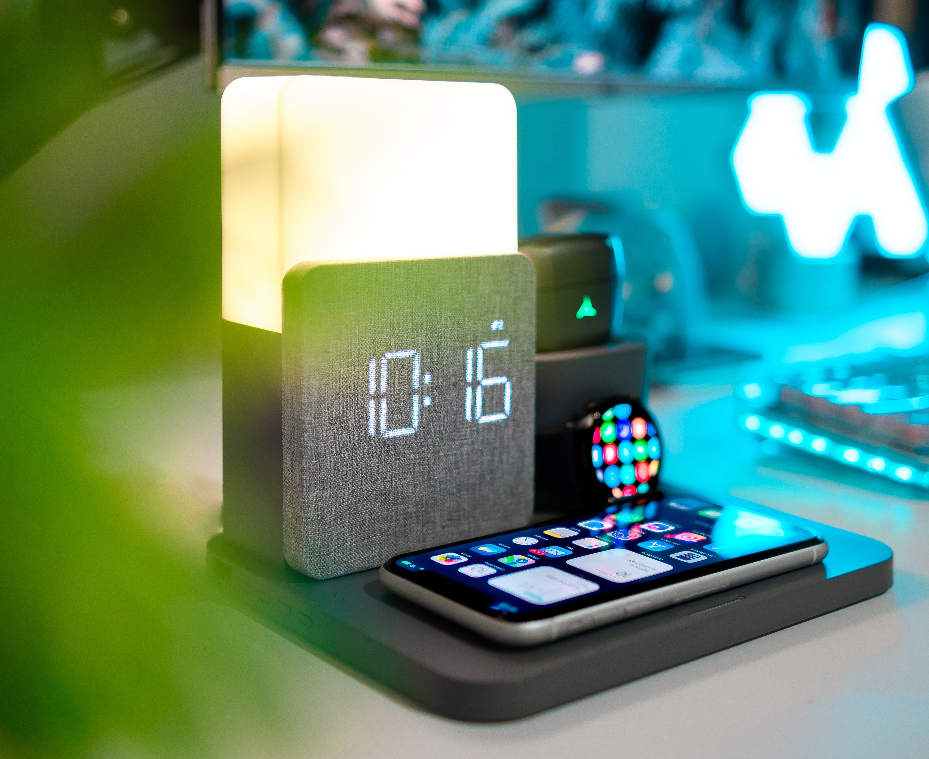 Dark grey alarm clock with wireless charging iphone, airpods and apple watch on a gaming desk with blue led lights in the back