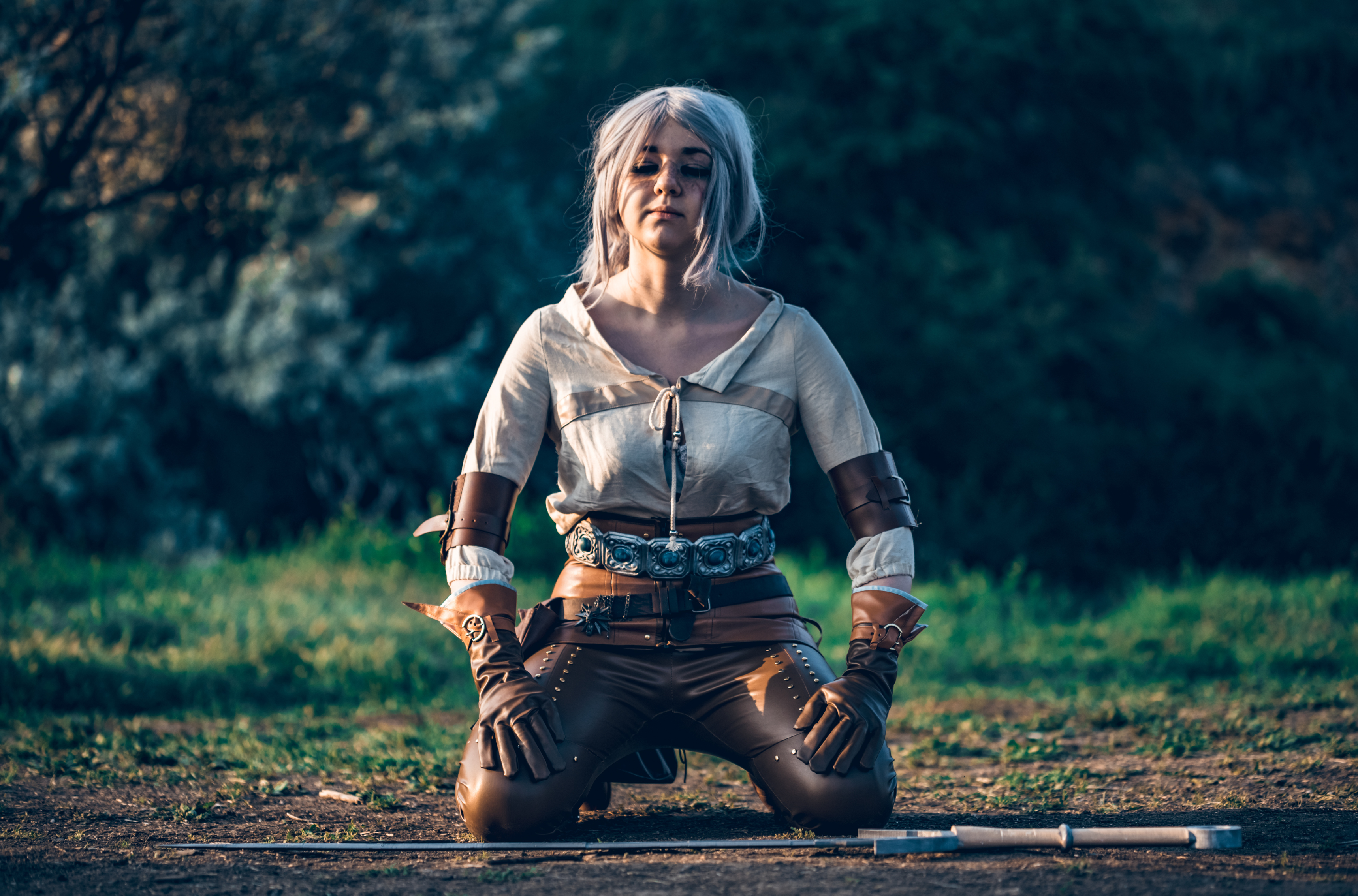 An image from The Witcher 3, depicting Ciri, a young female warrior with white hair, kneeling with her sword planted in the ground beside her, displaying a determined and focused expression in a dramatic and atmospheric setting